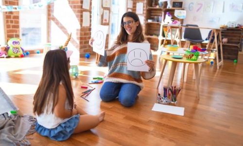 Beautiful pshychologist and toddler doing therapy with emotions emoji around lots of toys at kindergarten